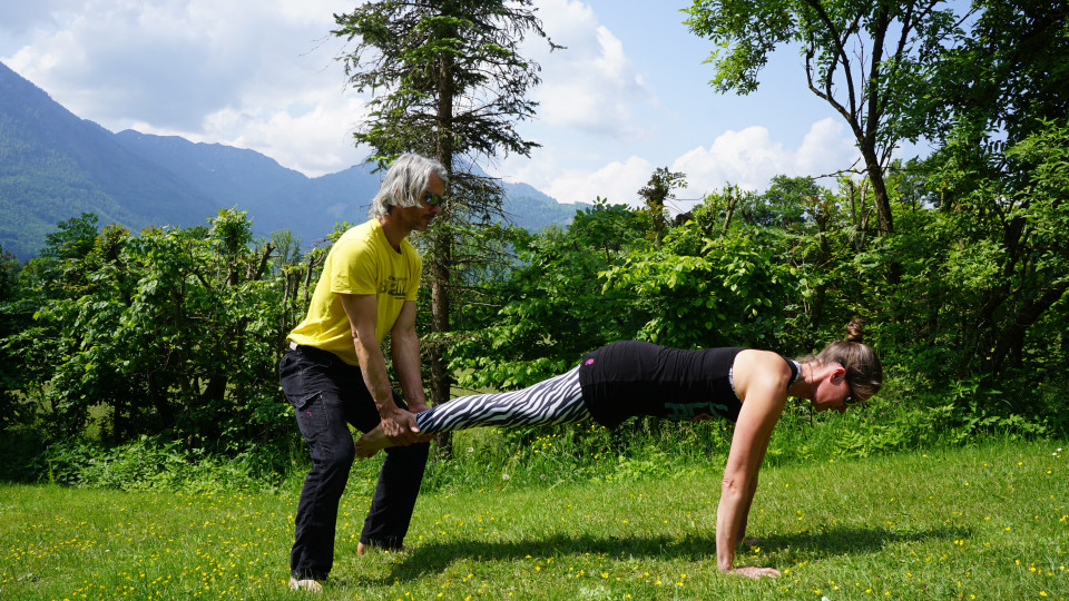 Wie im Plank Drill in die Brettposition kommen und ein Partner hält hinten die Fußgelenke. Die Person im Brett kommt nun hoch in eine L-Position und bringt das Becken über die Schultern. Der Partner wandert hinten mit. 